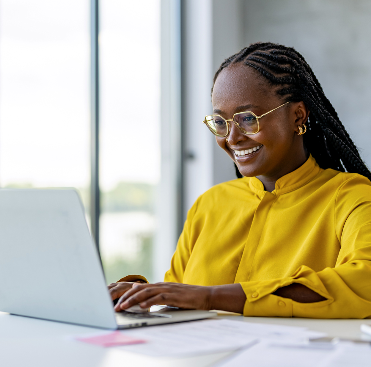 A focused woman wearing a yellow shirt and glasses, diligently working on answering customer questions from her laptop.