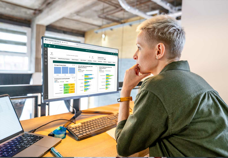 A woman wearing a green shirt is looking at a computer displaying a UKG product.