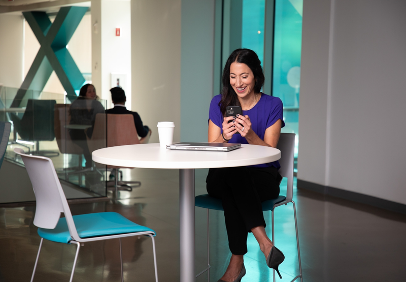 A young woman sits at a table in an office break room, smiling while looking at her phone.